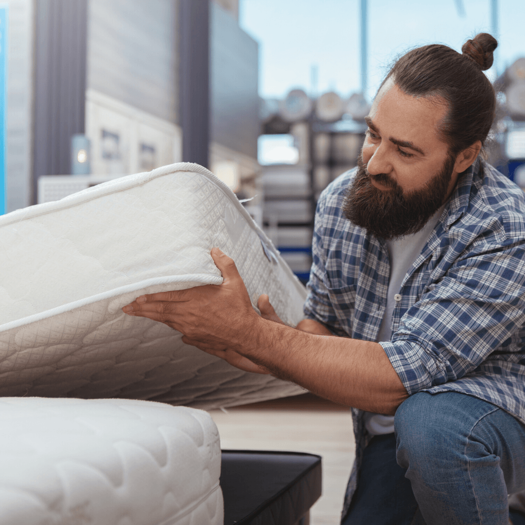 man in a bed shop crouching down by a mattress and lifting it up to look at more closely